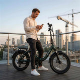 Zielony rower elektryczny DYU C9 na miejskim tarasie, obok którego stoi mężczyzna korzystający z telefonu, z panoramą miasta w tle.